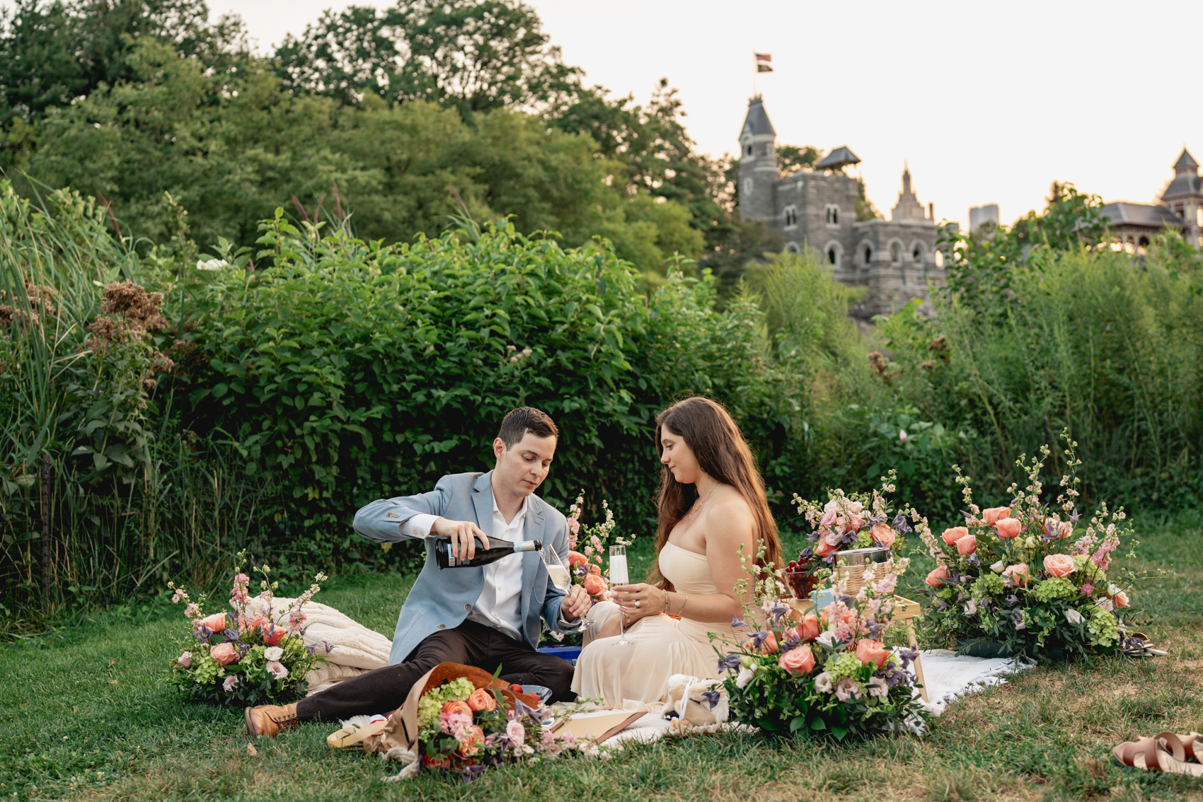 Couple sharing a drink on a flower-covered picnic blanket with Belvedere Castle rising behind the trees in Central Park