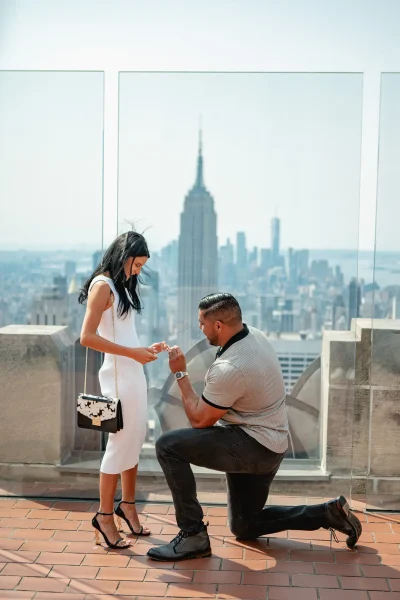 Man proposing at Top of the Rock in New York City as his partner admires the ring. The Empire State Building stands prominently in the background, making this rooftop engagement moment iconic and unforgettable. Perfect photo for planning your Top of the Rock proposal at Rockefeller Center.
