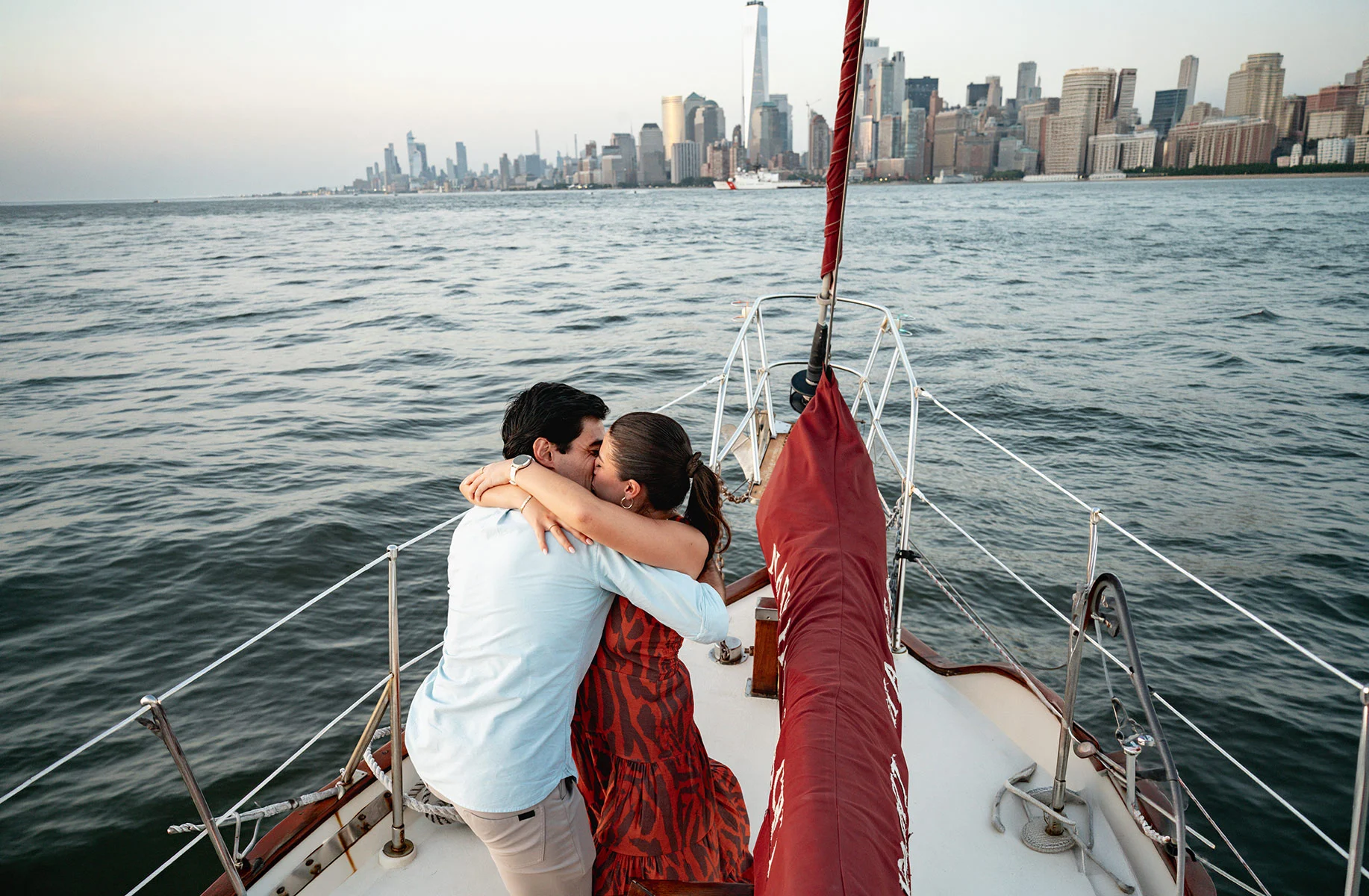 Couple hugging on the bow of a sailboat with a red sail and the distant Manhattan skyline over the water