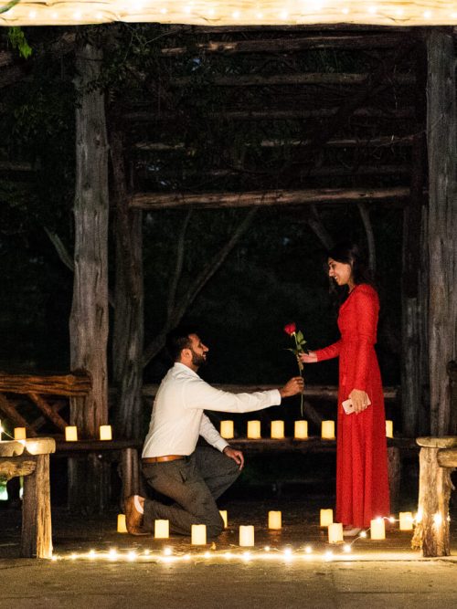 Man kneeling with a red rose to a woman in a red dress inside a candlelit rustic gazebo at night