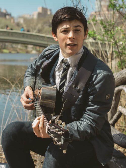 Young man in a leather jacket singing and playing guitar by the lake with Bow Bridge behind him in Central Park
