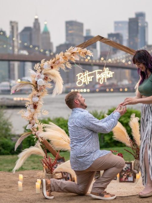 Man kneeling to propose under a pampas grass arch with a Better Together neon sign near the Brooklyn Bridge