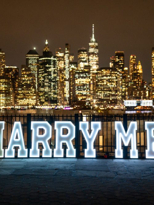 Four-foot illuminated MARRY ME letters on a waterfront deck facing the Manhattan skyline at night