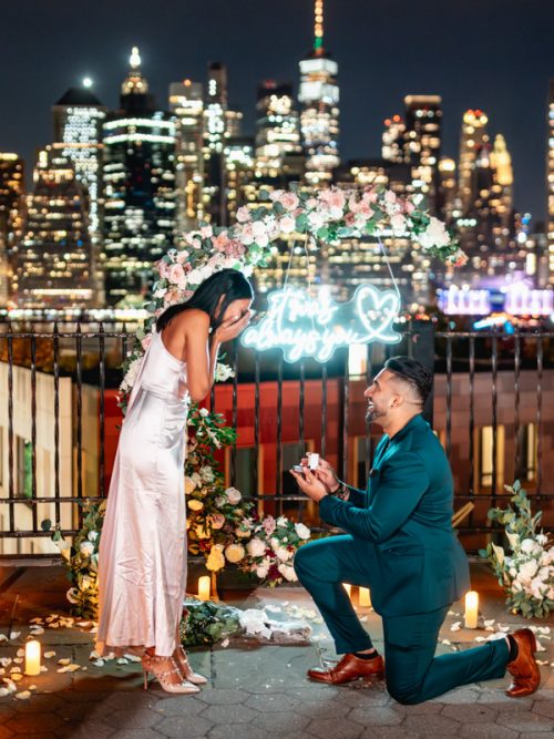 Man in a teal suit kneeling before a woman in a pink satin dress beside a floral arch, candles and the skyline glowing