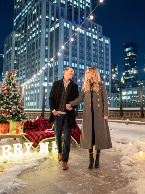 Couple in long coats walk past a Christmas tree, Marry Me letters and candles on a snowy NYC rooftop at night