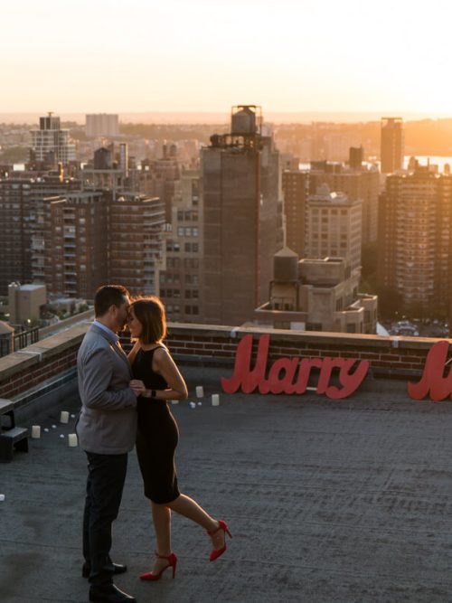 Couple embracing beside red Marry Me letters and scattered candles on a Manhattan rooftop at sunset