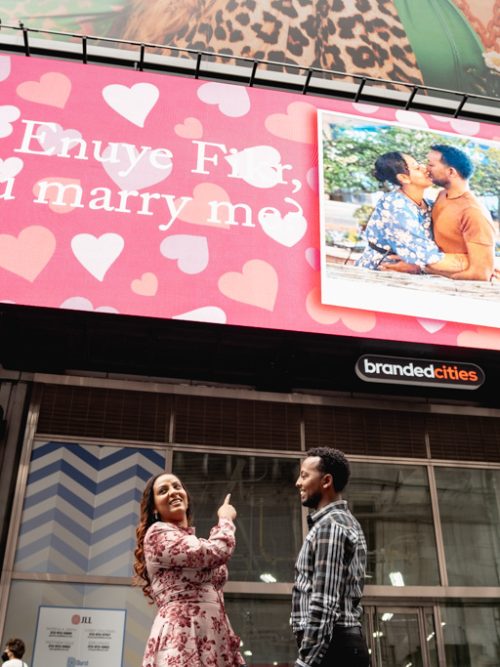 Couple beneath a pink heart-filled Times Square billboard displaying a Will you marry me message and a couple's photo