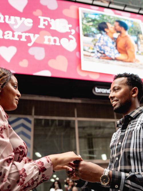 Couple holding hands in Times Square below a pink billboard with hearts reading Will you marry me