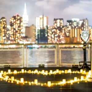 Glowing candle heart on a waterfront pier at night with a blurred Manhattan skyline and the Empire State Building beyond