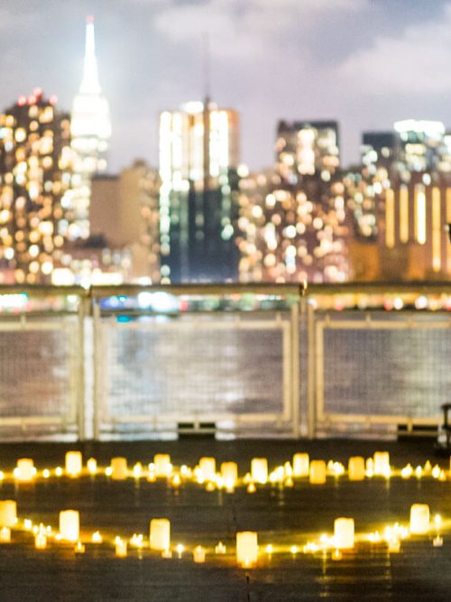 Glowing candle heart on a waterfront pier at night with a blurred Manhattan skyline and the Empire State Building beyond