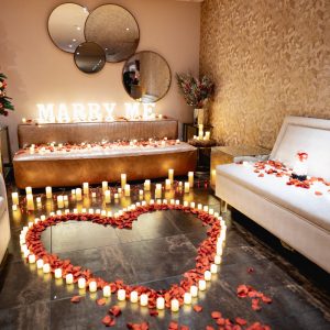 Hotel room with a glowing Marry Me sign, a heart of candles and red rose petals, a small Christmas tree and mirrors