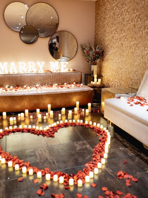 Hotel room with a glowing Marry Me sign, a heart of candles and red rose petals, a small Christmas tree and mirrors