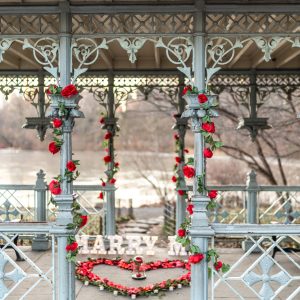 Central Park cast-iron gazebo with red roses on the columns, MARRY ME letters and a rose petal heart on the floor
