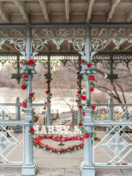Central Park cast-iron gazebo with red roses on the columns, MARRY ME letters and a rose petal heart on the floor