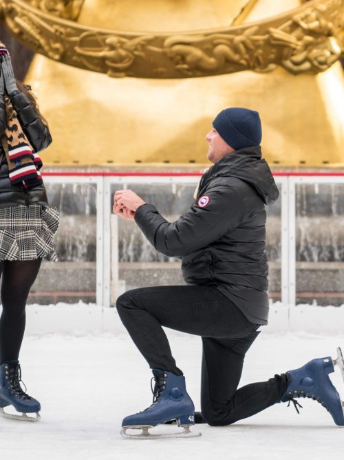 Man kneels on skates as a woman covers her face on an ice rink in front of a golden statue