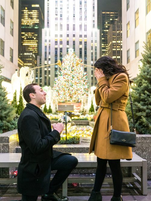 Man kneels with a ring before a woman covering her face in front of the lit Rockefeller Center Christmas tree at dusk