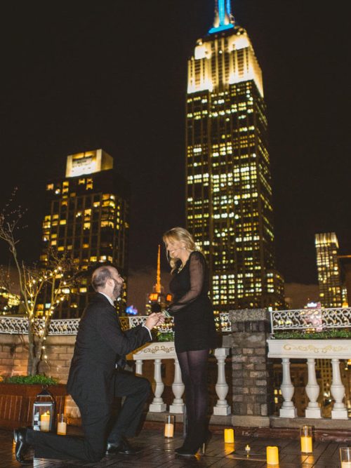 Man kneeling to a woman in a black dress on a candlelit stone-balustrade rooftop with the Empire State Building lit at night