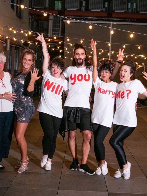 Group of cheering dancers in white Will You Marry Me? T-shirts pose on a rooftop terrace under string lights at night