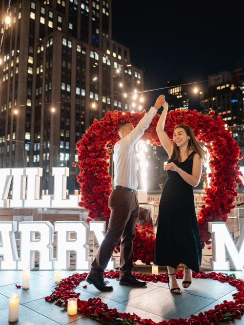Couple twirls beneath a red rose heart arch beside lit WILL YOU MARRY ME letters on a string-lit Manhattan rooftop at night