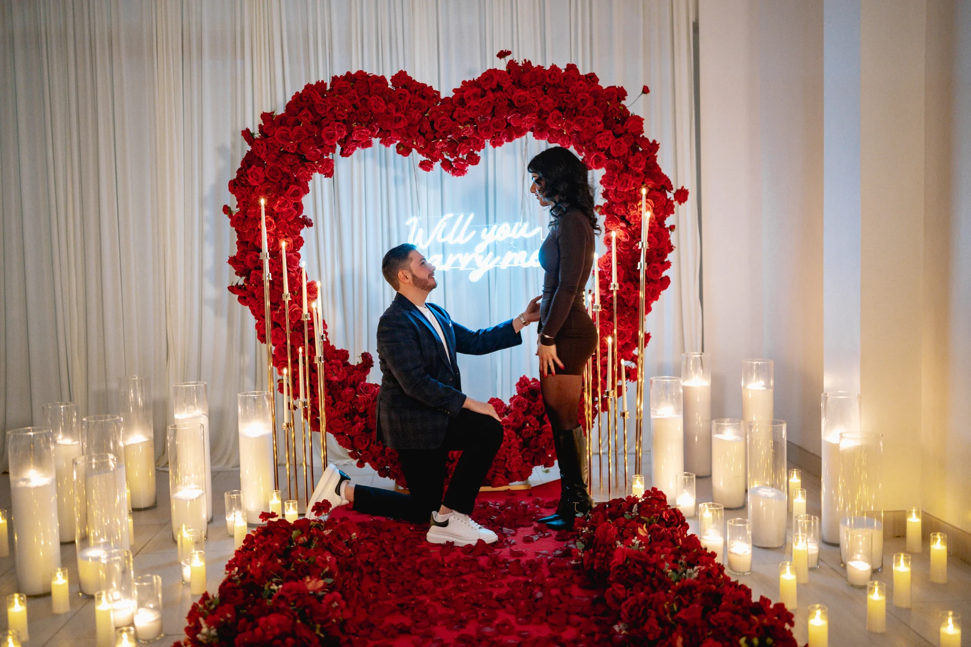 Man kneeling before his partner in front of a giant red rose heart with a Will You Marry Me neon, surrounded by candles