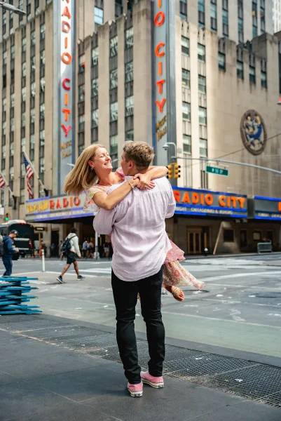 Joyful moment in front of Radio City Music Hall as the couple celebrates their Top of the Rock engagement at Rockefeller Center.