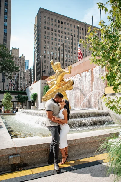 Couple celebrating their engagement with a kiss at the Rockefeller Center fountain, near the golden Prometheus statue, after a Top of the Rock proposal.