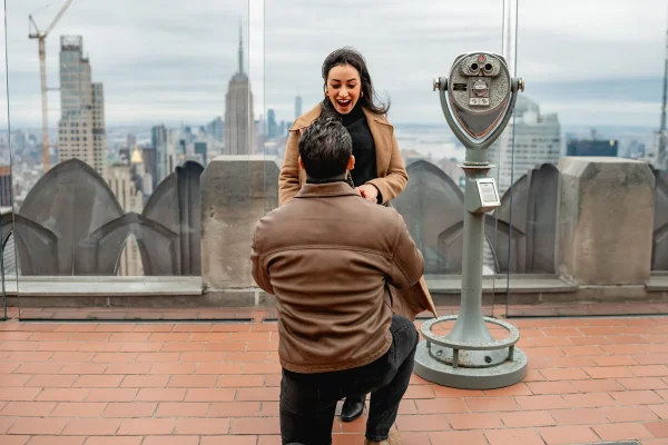 Surprise proposal captured at Top of the Rock in Rockefeller Center with skyline view and joyful reaction.