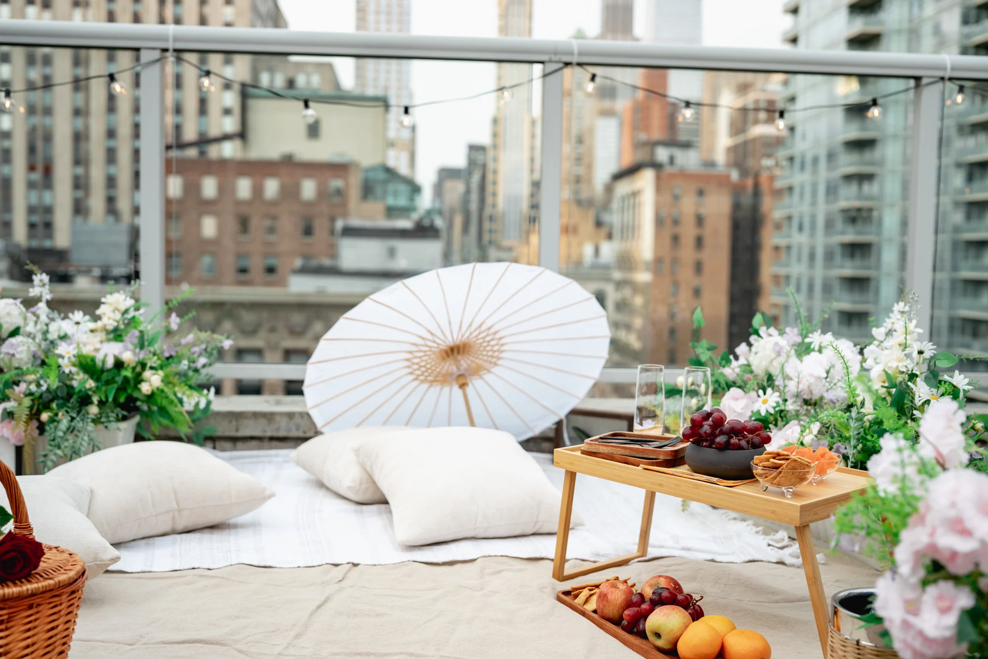 White paper parasol, pillows and wooden tray on a picnic blanket by a glass railing with string lights