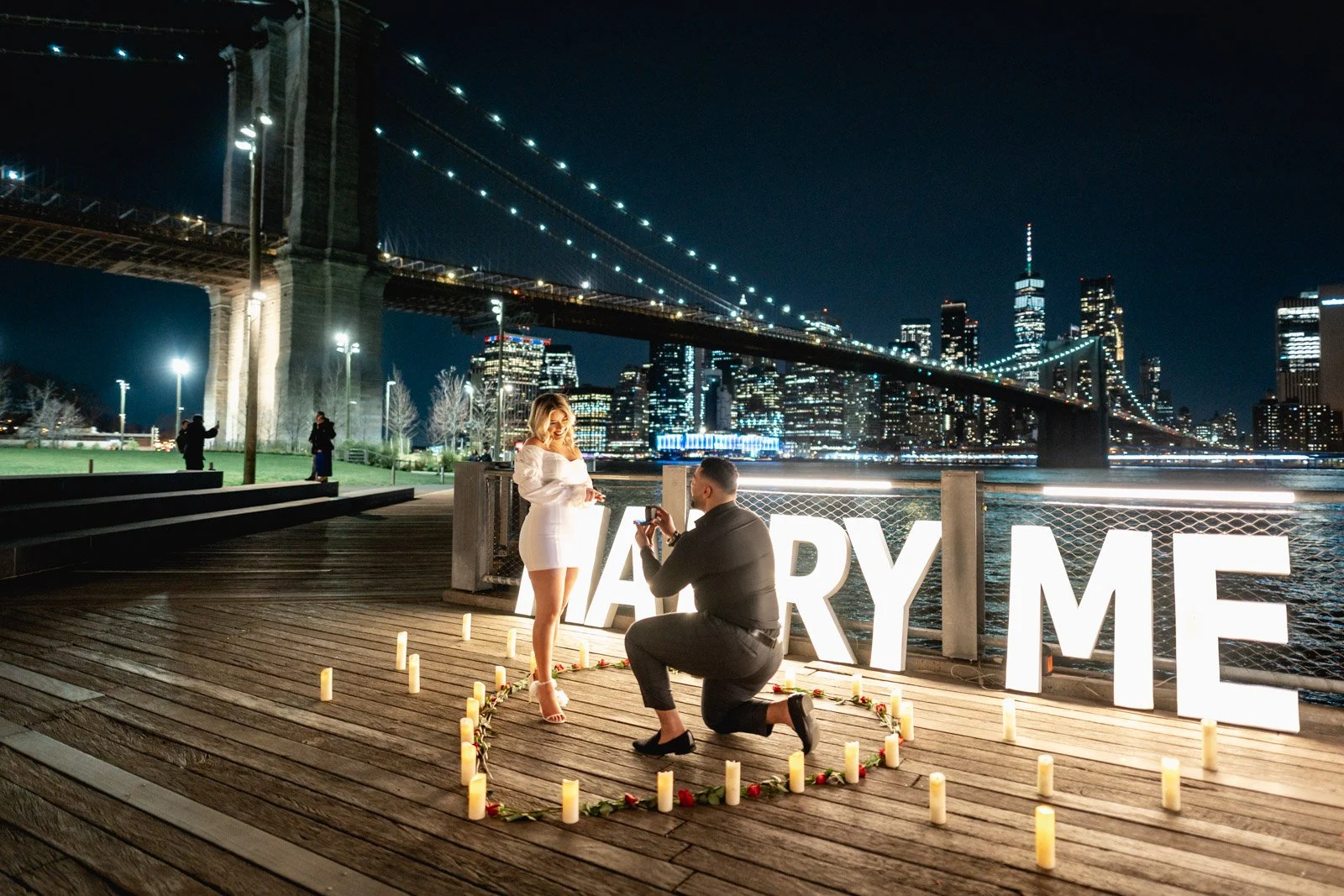 Man kneels in a candle and rose heart before a woman in white at giant MARRY ME letters beneath the Brooklyn Bridge