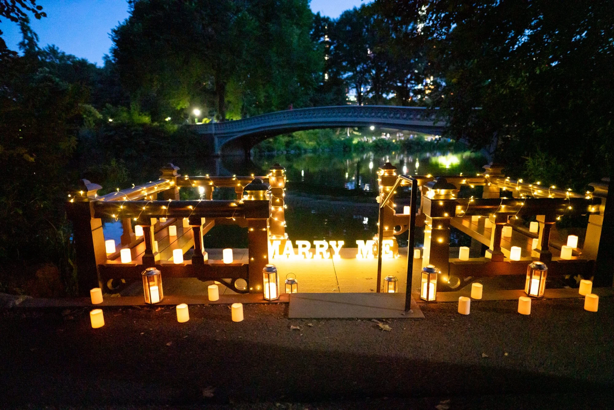 Candlelit wooden landing with MARRY ME letters by Bow Bridge in Central Park at dusk