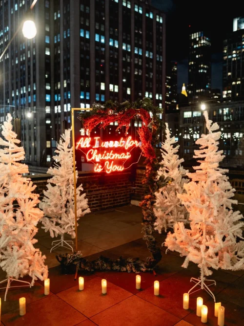Red neon All I Want for Christmas is You sign in an arch between white Christmas trees on a rooftop at night