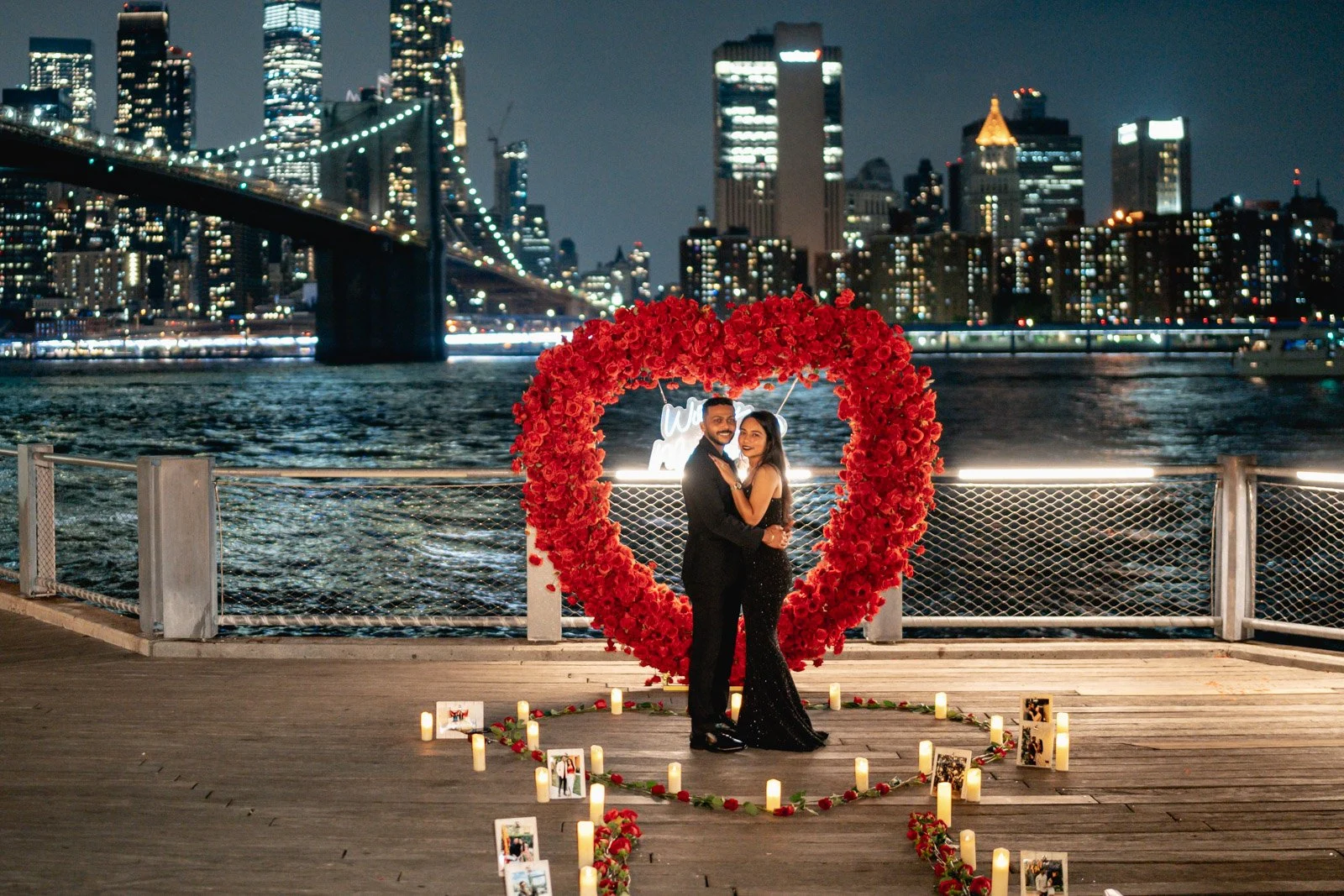 Couple embracing inside a red rose heart arch on a waterfront deck with the Brooklyn Bridge lit at night