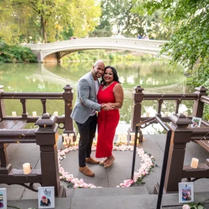Couple standing close in front of a pink rose heart and candles on a lakeside landing, with Bow Bridge across the water