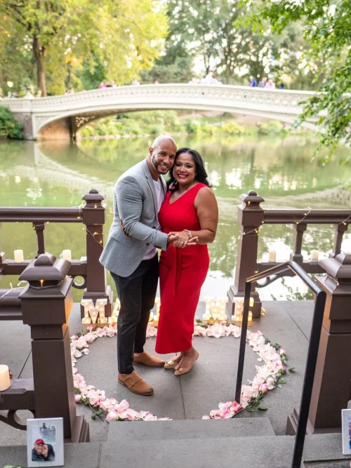 Couple standing close in front of a pink rose heart and candles on a lakeside landing, with Bow Bridge across the water