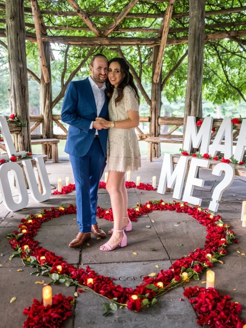 Couple standing in a red rose petal heart between giant WILL YOU MARRY ME letters in a rustic Central Park gazebo