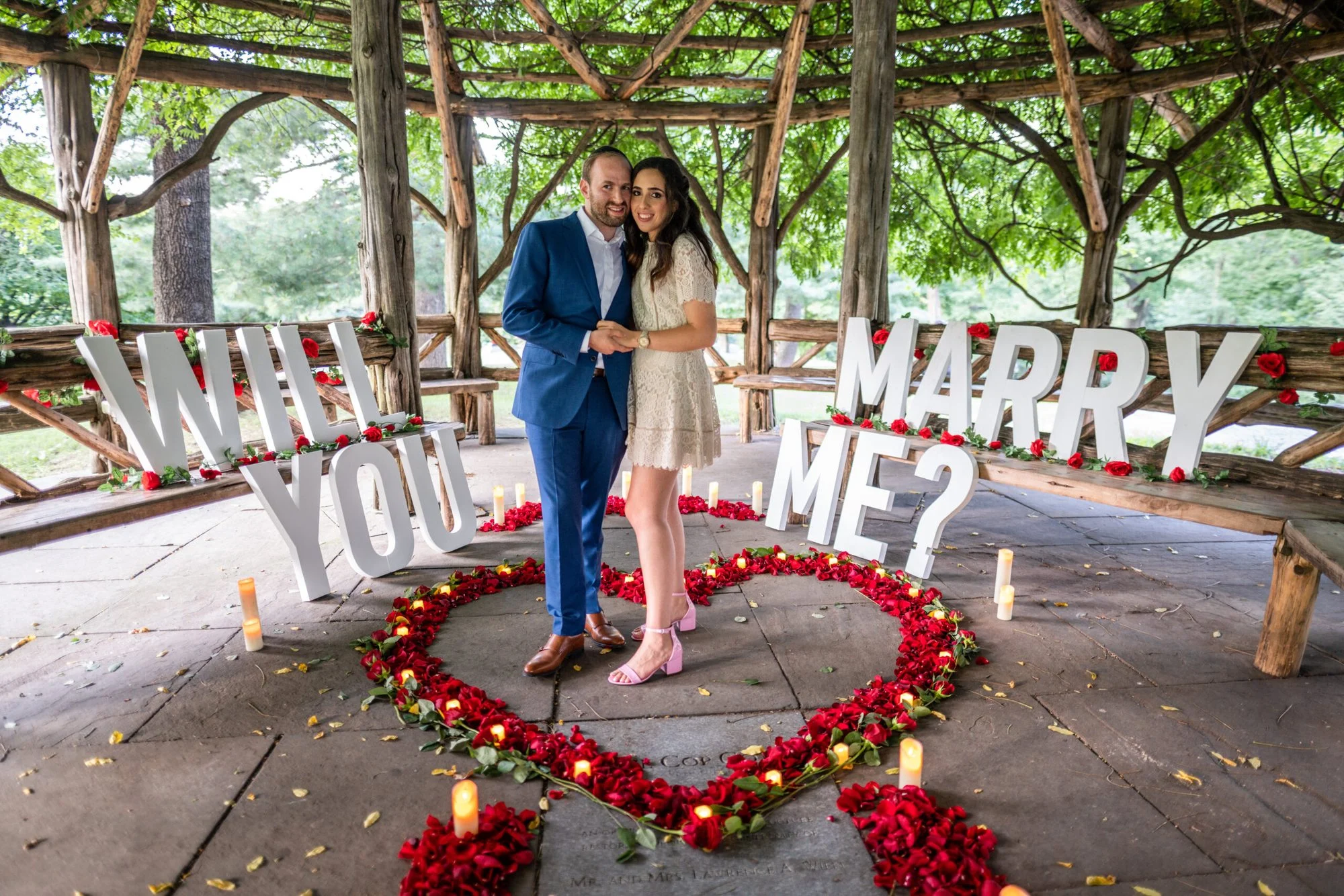 Couple standing in a red rose petal heart between giant WILL YOU MARRY ME letters in a rustic Central Park gazebo
