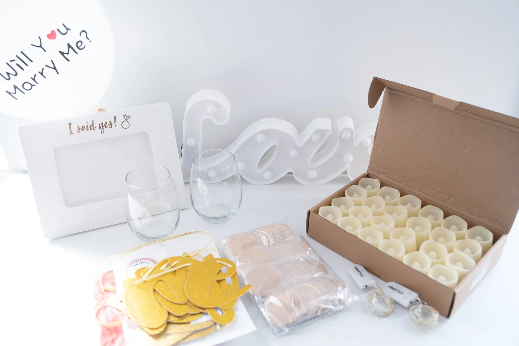 Flat lay of a DIY proposal kit with a Love marquee sign, etched glasses, candles, gold hearts and a Will You Marry Me balloon