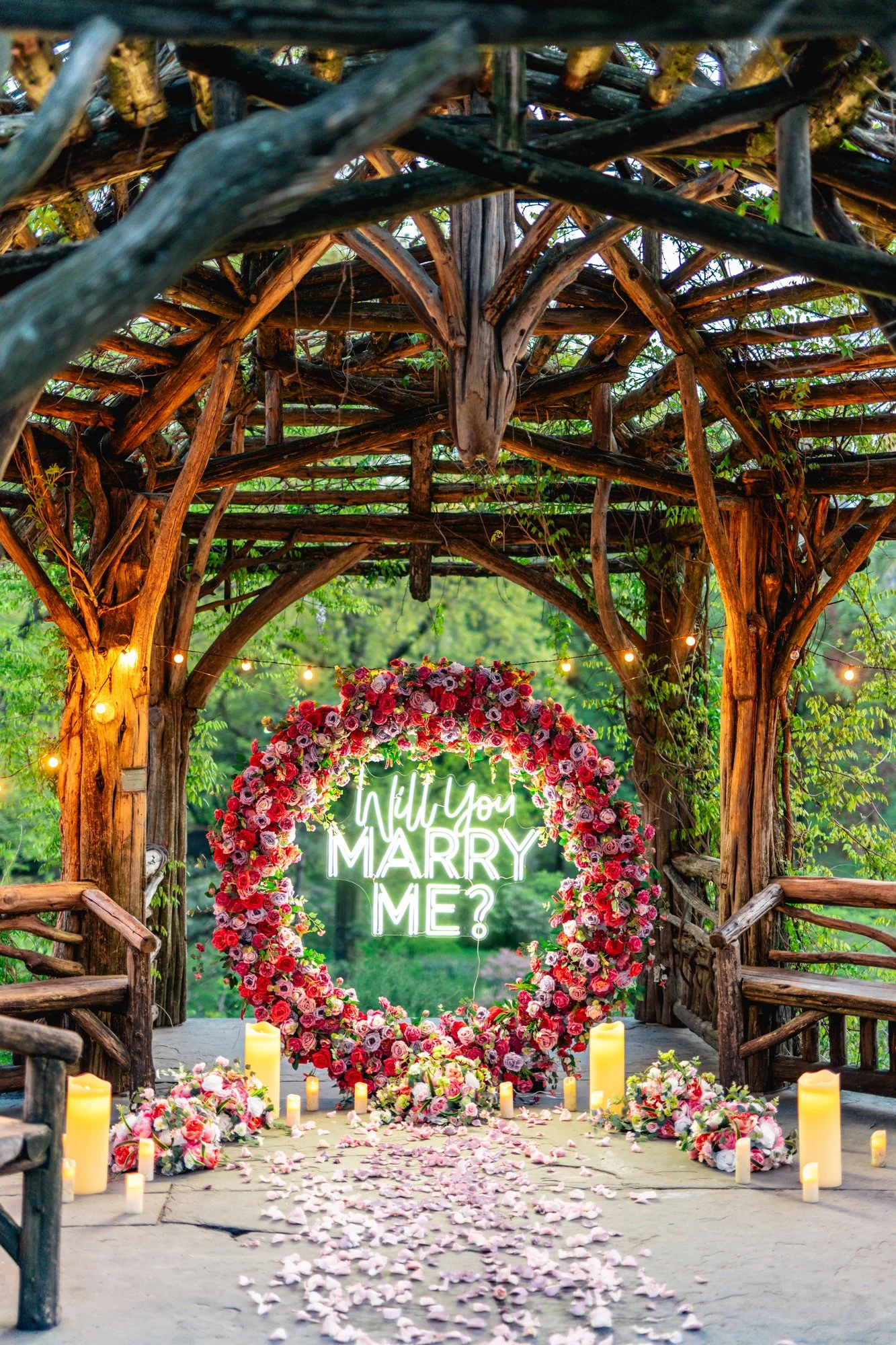 Round red and pink flower arch with a Will You Marry Me neon sign, candles and petals in a rustic Central Park gazebo