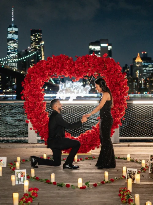 Man kneeling before a woman in a black gown inside a red rose heart arch by the water with the Brooklyn Bridge glowing