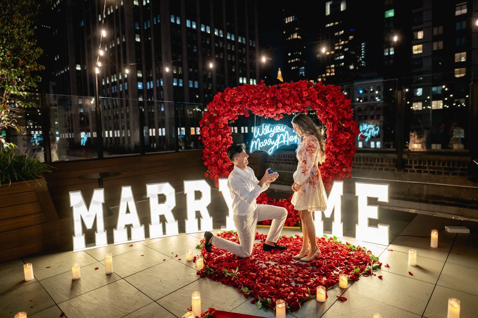 Man kneeling before a woman in a rose petal heart beneath a red rose heart arch and lit MARRY ME letters at night