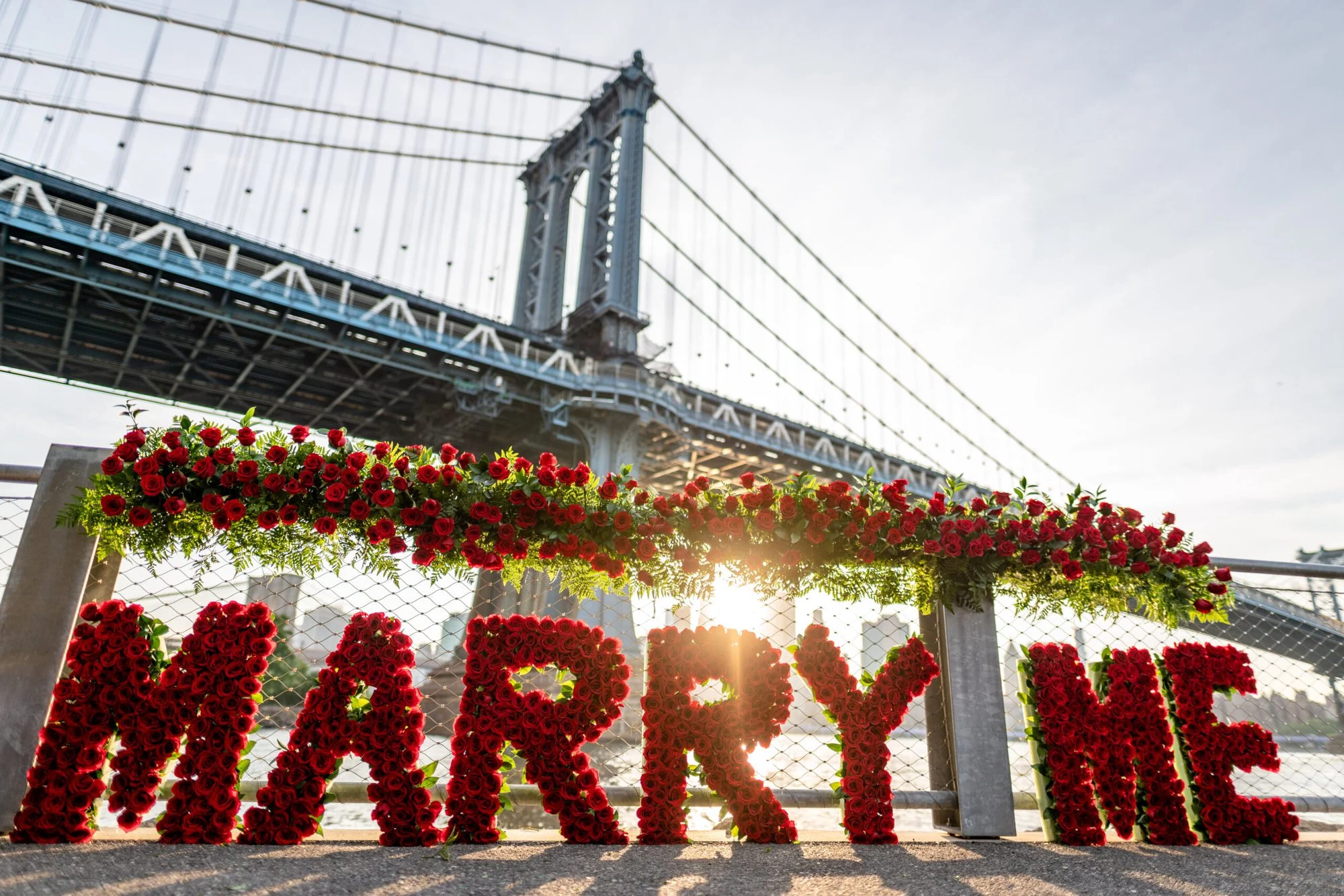 Large red rose letters spelling MARRY ME under a garland of roses, with the Manhattan Bridge and sun glow behind