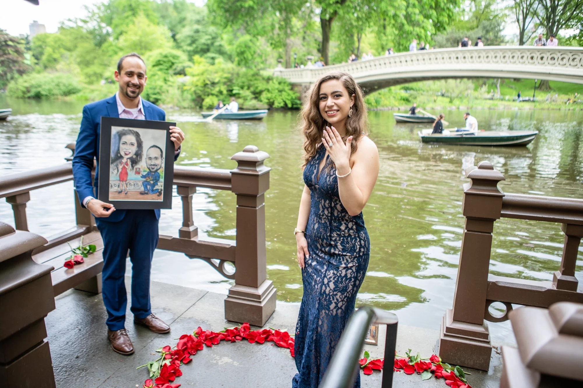 Man in a blue suit holds a caricature portrait beside a woman showing her ring, with red rose petals and Bow Bridge behind