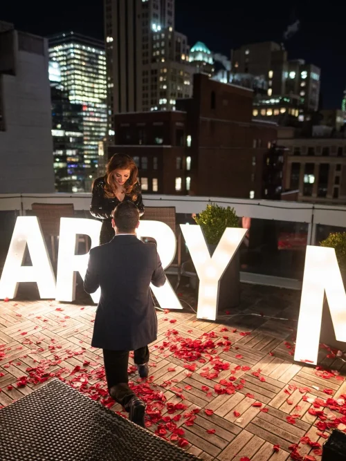 Man in a dark suit walks toward a woman behind giant illuminated MARRY ME letters on a petal-covered rooftop at night