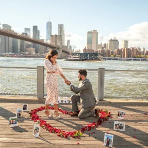 Man in a gray suit kneels inside a red rose petal heart with framed photos on a Dumbo pier, skyline behind