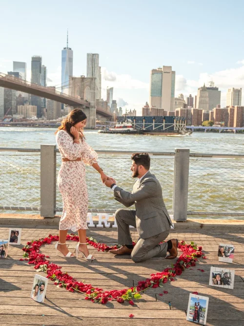 Man in a gray suit kneels inside a red rose petal heart with framed photos on a Dumbo pier, skyline behind