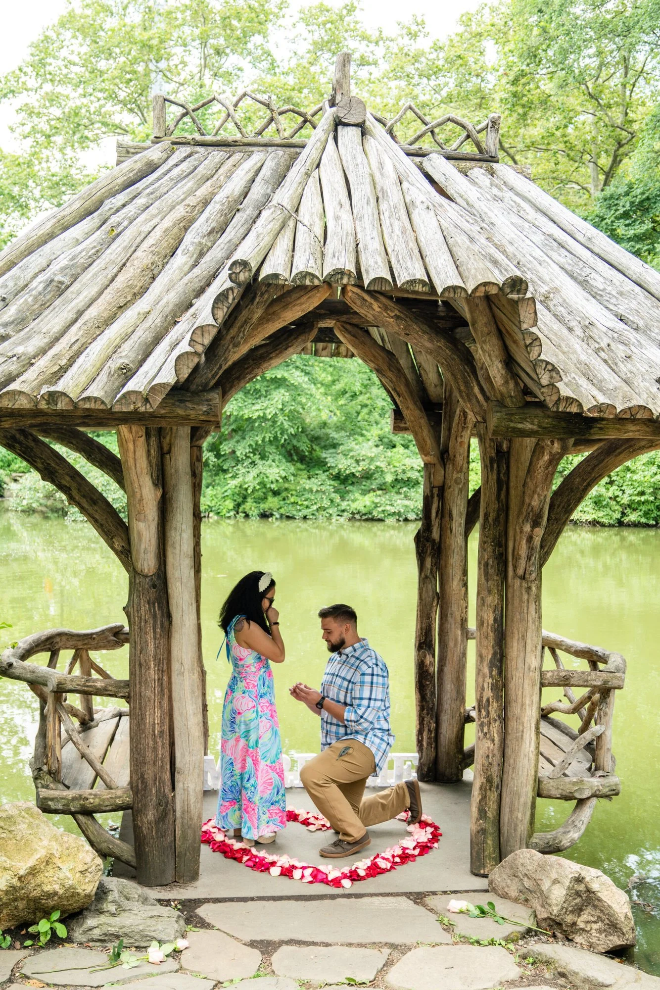 Man kneeling inside a rustic log gazebo above a green lake, with a rose-petal heart and his partner covering her face