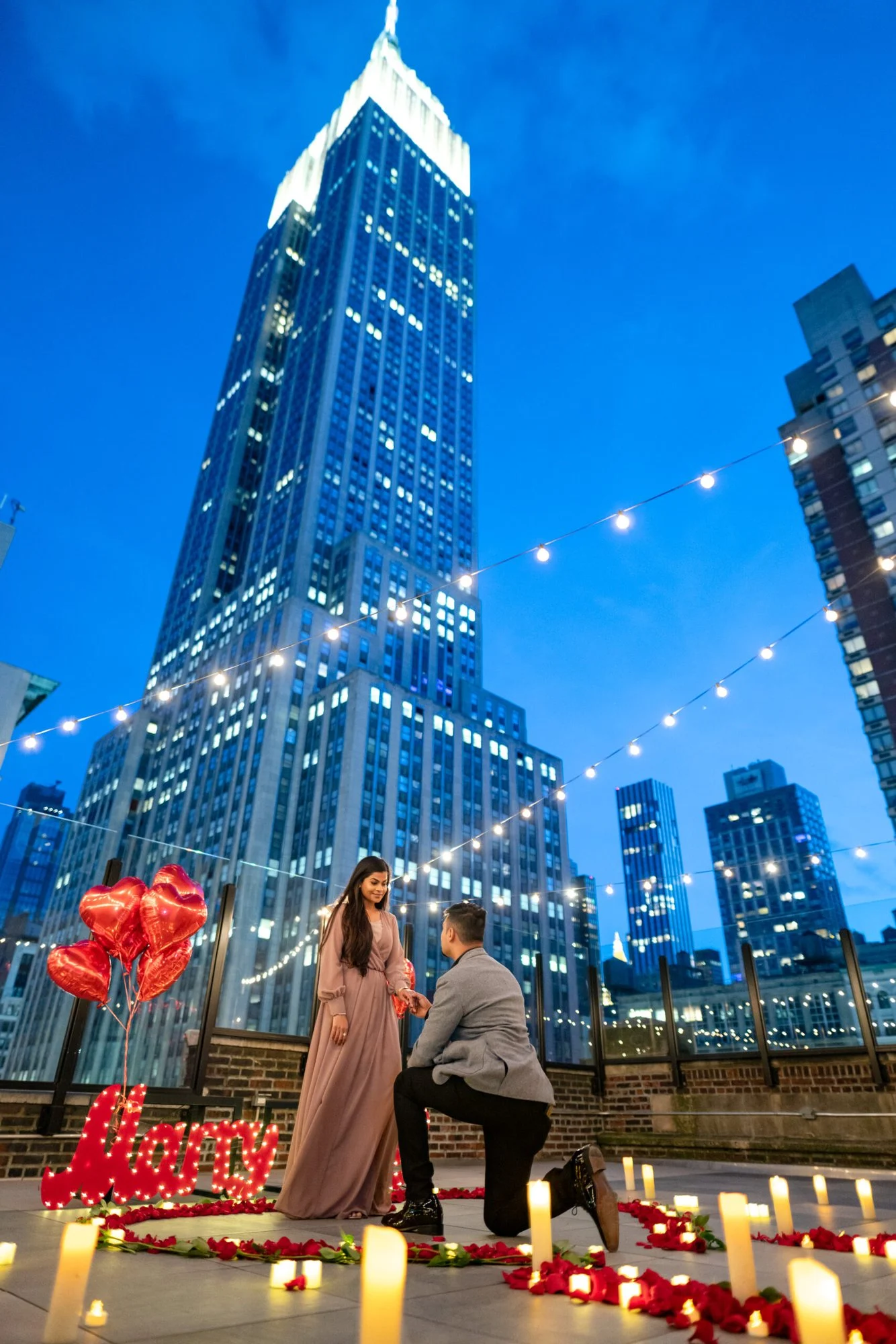 Man kneeling before a woman on a rooftop at blue hour with heart balloons, roses and the lit Empire State Building behind