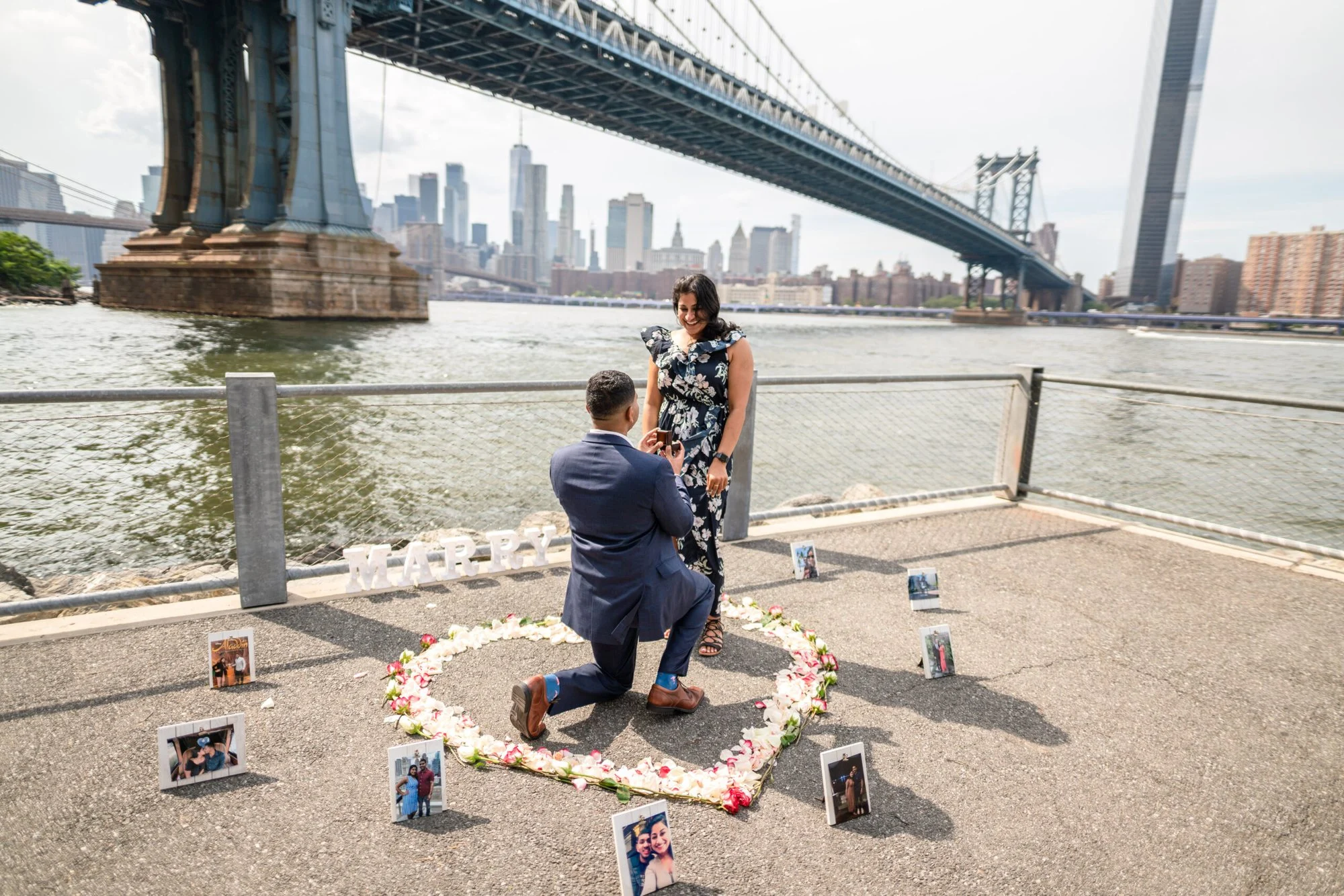 Man kneels in a white flower heart ringed with framed photos on a Dumbo pier beneath the Manhattan Bridge