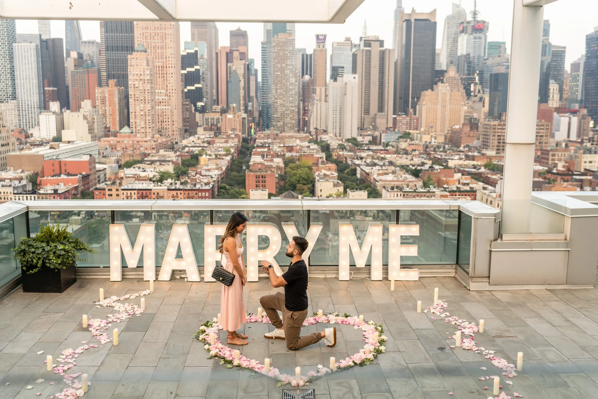 Man kneels before a woman in a pink dress inside a rose-petal heart at MARRY ME letters with Midtown rooftops behind