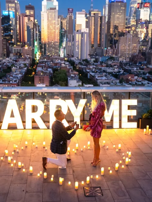 Man kneels before a woman in a pink dress at lit MARRY ME letters inside a candle heart, Manhattan skyline at dusk